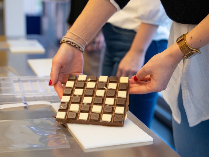 Person h&auml;lt eine Schokoladentafel mit Schachbrettmuster in einer Werkstatt. Im Hintergrund sind weitere Personen und Materialien zu sehen., &copy; Alfred Ritter GmbH &amp; Co. KG | Waldenbuch