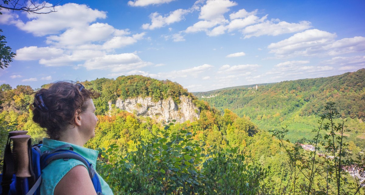 Eine Frau mit Rucksack blickt auf eine bewaldete Felslandschaft, den Geiselstein, unter einem blauen Himmel mit Wolken., © Stadt Geislingen an der Steige Eine Frau mit Rucksack blickt auf eine bewaldete Felslandschaft, den Geiselstein, unter einem blauen Himmel mit Wolken., © Stadt Geislingen an der Steige