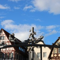 Das Schäferlauf-Denkmal zeigt Figuren und Glocken auf einem Bogen vor Fachwerkhäusern unter blauem Himmel., © Bad Urach Tourismus