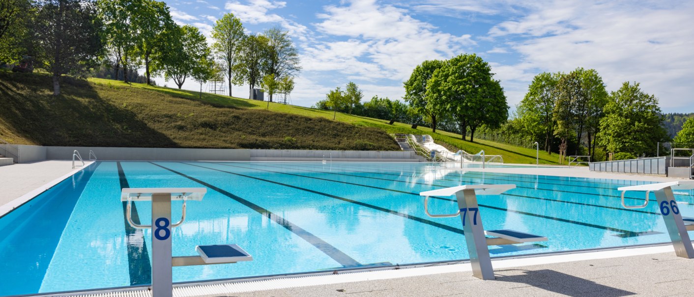 Ein leeres Freibad mit Startblöcken und einer Wasserrutsche, umgeben von grünen Bäumen und blauem Himmel., © Stadt Gaildorf Ein leeres Freibad mit Startblöcken und einer Wasserrutsche, umgeben von grünen Bäumen und blauem Himmel., © Stadt Gaildorf