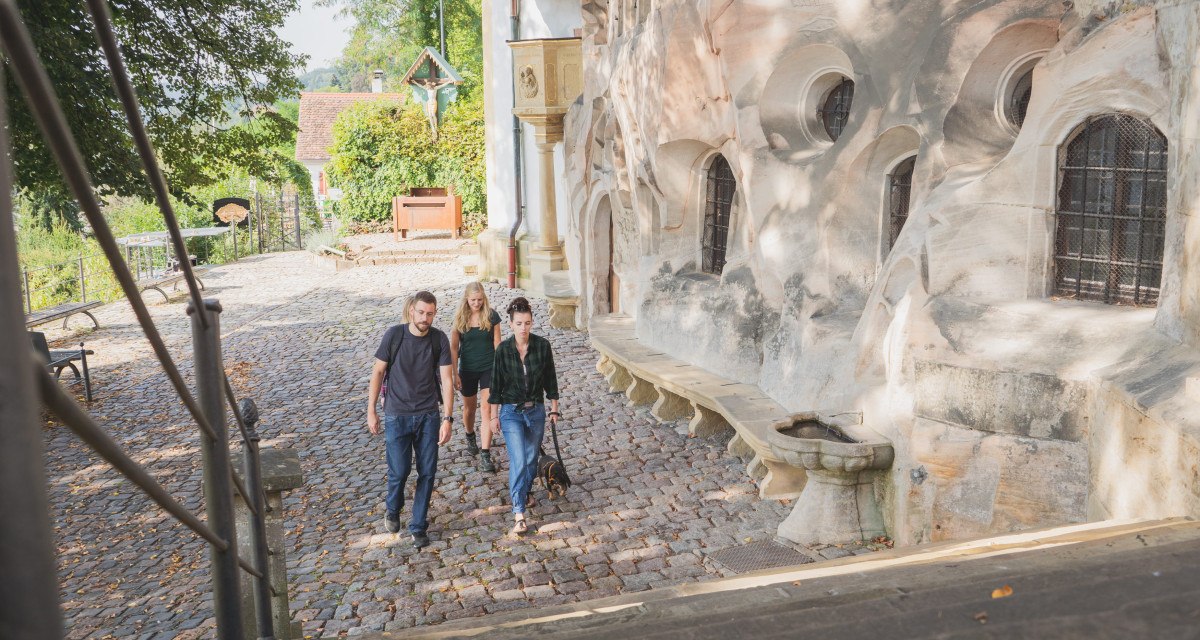 Vier Personen spazieren auf einem gepflasterten Weg neben einer historischen Steinmauer in Schw&auml;bisch Gm&uuml;nd. Im Hintergrund sind B&auml;ume und ein kleines Geb&auml;ude zu sehen.