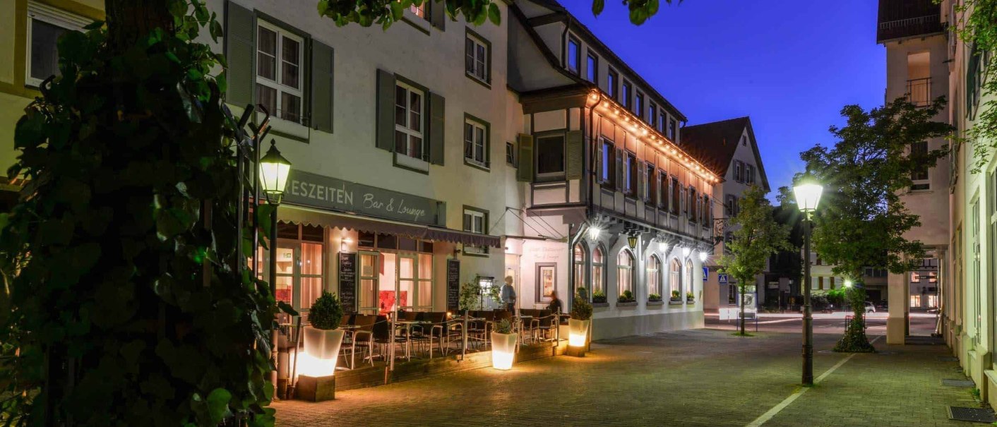 Ein beleuchtetes Hotelgeb&auml;ude bei Nacht, umgeben von B&auml;umen und Stra&szlig;enlaternen. Vor dem Hotel stehen Tische und St&uuml;hle., &copy; Flairhotel Vier Jahreszeiten