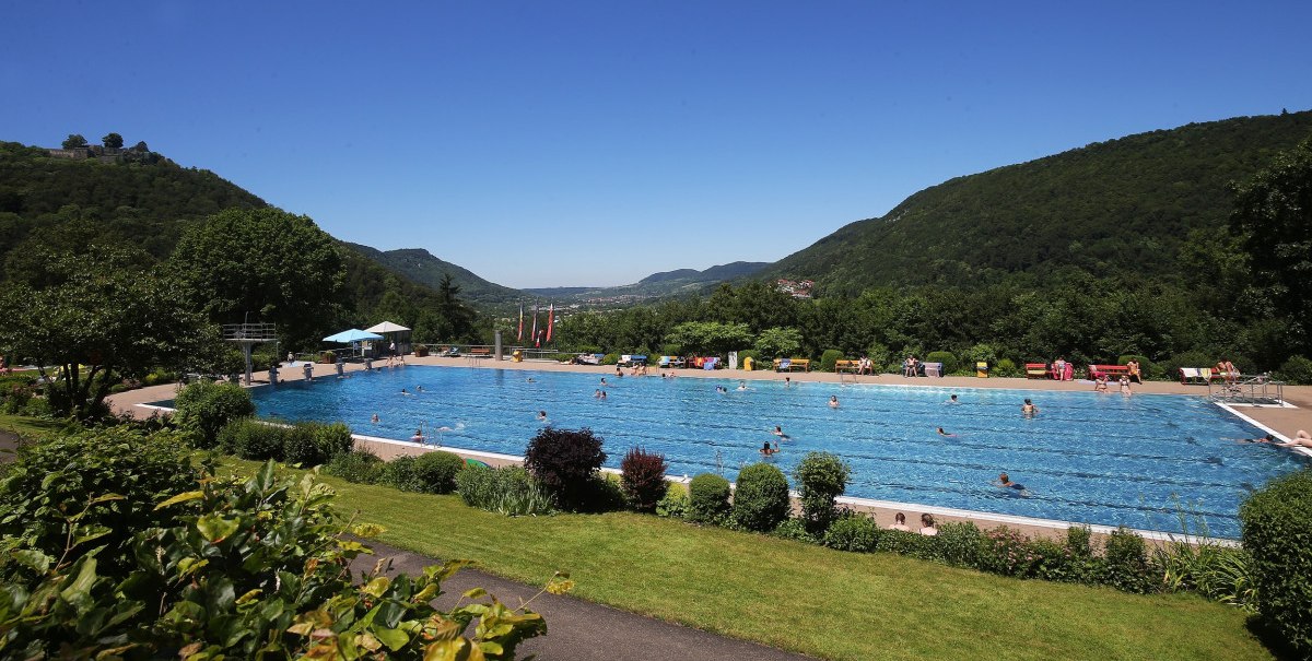 Ein Freibad inmitten einer gr&uuml;nen, bergigen Landschaft. Menschen schwimmen im gro&szlig;en Becken, umgeben von B&auml;umen und H&uuml;geln unter klarem Himmel.