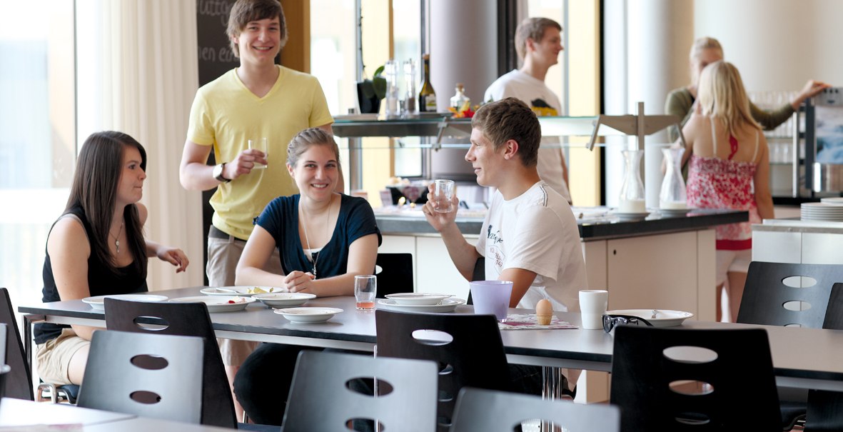 Junge Leute sitzen und stehen in einem modernen Speisesaal, unterhalten sich und trinken. Im Hintergrund ist eine Theke mit Geschirr zu sehen., © DJH Landesverband Baden-Württemberg e.V.