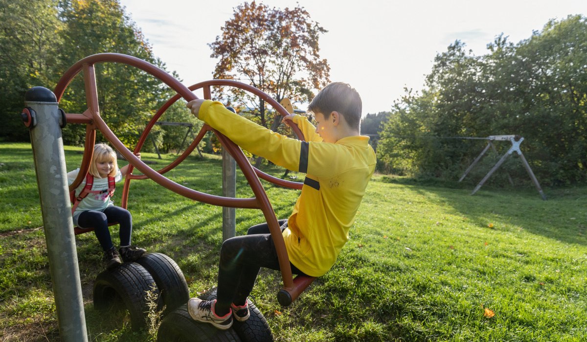 Zwei Kinder spielen auf einem Klettergerüst aus Reifen und Metallstangen auf einem grünen Spielplatz. Im Hintergrund sind Bäume und eine Schaukel zu sehen., © Foto Thomas Zehnder