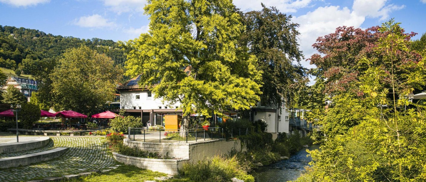 Ein grüner Park mit Bäumen, einem kleinen Fluss und einem Gebäude mit roten Sonnenschirmen. Im Hintergrund sind Hügel zu sehen., © Stuttgart-Marketing GmbH, Sarah Schmid