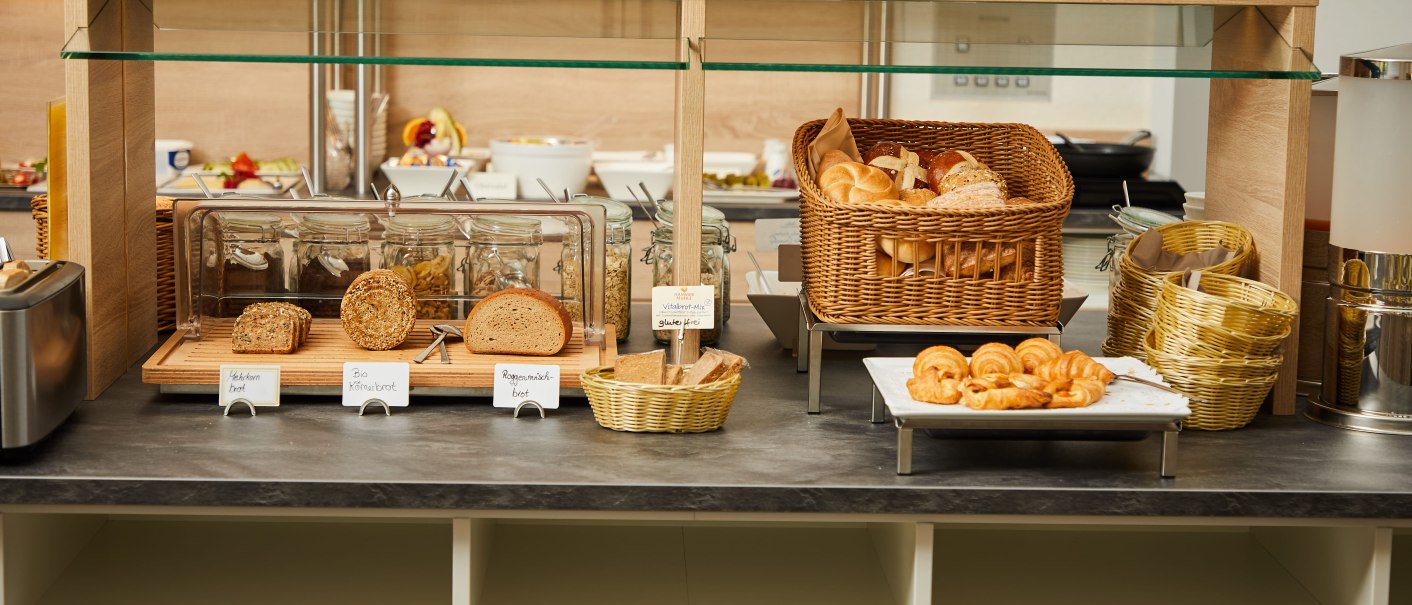 Ein Frühstücksbuffet mit verschiedenen Brotsorten, Brötchen und Croissants. Gläser und Körbe sind ebenfalls zu sehen., © TOMAS