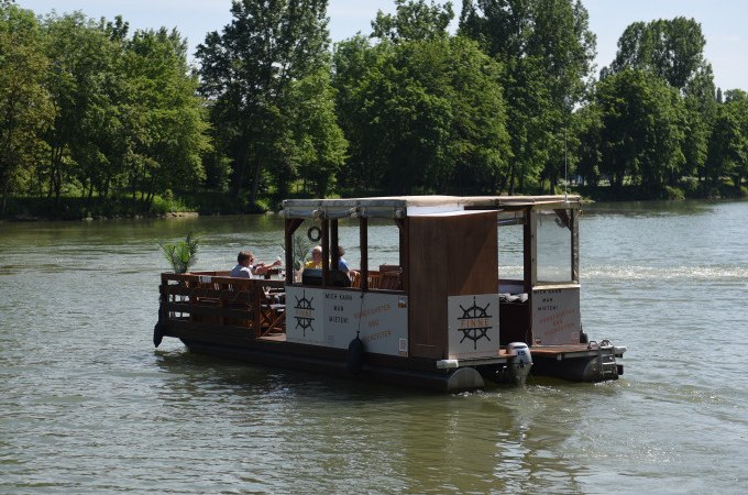 Ein rustikales Floßboot mit überdachtem Bereich fährt auf einem Fluss. Im Hintergrund sind grüne Bäume zu sehen., © Cool-Tours StattReisen