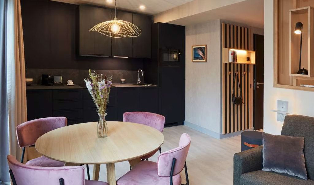 Modernes Apartment mit rundem Holztisch, rosa Stühlen, dunkler Küche und stilvoller Beleuchtung. Gemütliche Atmosphäre mit Sofa und Dekoration., © Adagio Modernes Apartment mit rundem Holztisch, rosa Stühlen, dunkler Küche und stilvoller Beleuchtung. Gemütliche Atmosphäre mit Sofa und Dekoration., © Adagio