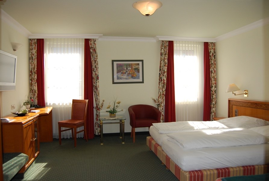 Helles Hotelzimmer mit Doppelbett, Schreibtisch, Stuhl, Sessel und dekorativen Vorhängen. Ein Bild hängt an der Wand., © Jeanette Bender Helles Hotelzimmer mit Doppelbett, Schreibtisch, Stuhl, Sessel und dekorativen Vorhängen. Ein Bild hängt an der Wand., © Jeanette Bender