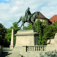 Reiterstatue auf dem Karlsplatz in Stuttgart, flankiert von L&ouml;wenstatuen und Obelisken, umgeben von B&auml;umen und einem historischen Geb&auml;ude im Hintergrund., &copy; Stuttgart-Marketing GmbH