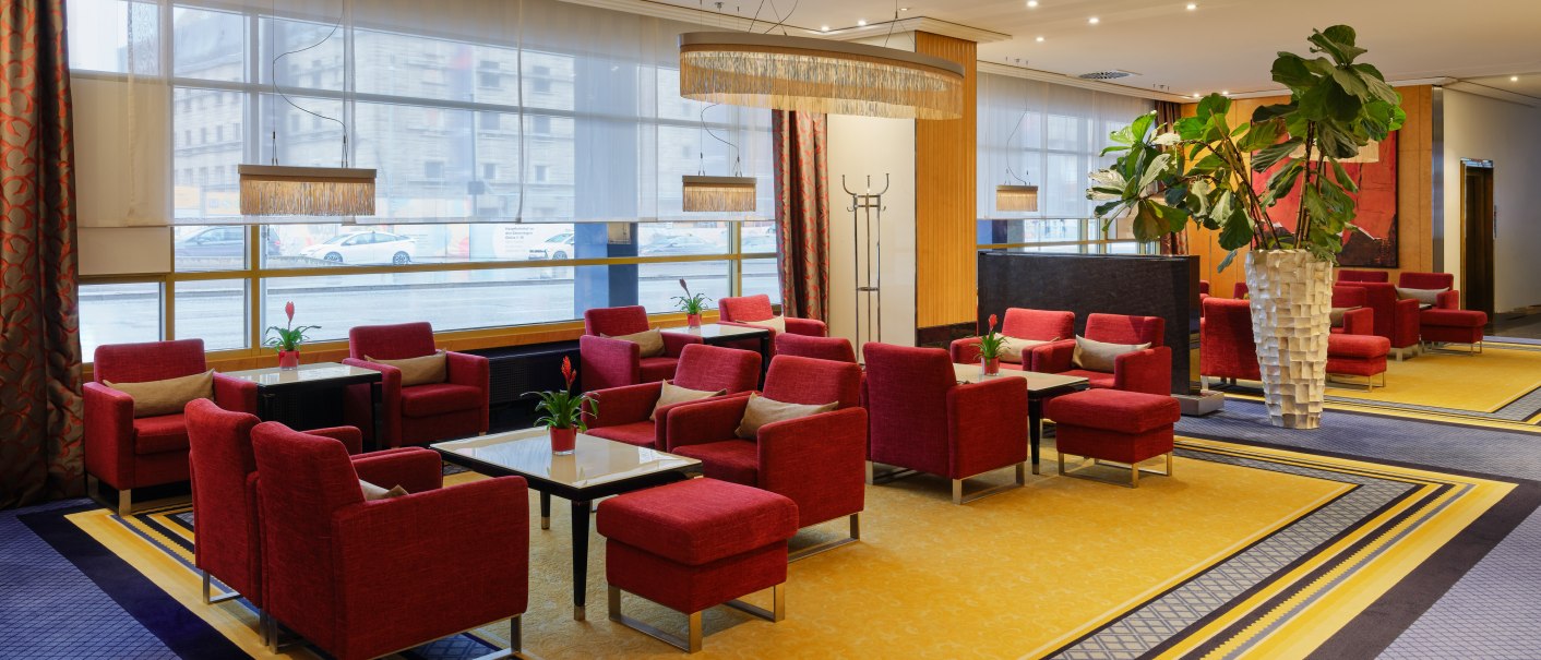 Elegante Lobby-Bar mit roten Sesseln, gelbem Teppich und großen Pflanzen. Große Fenster lassen viel Licht herein., © Steigenberger Graf Zeppelin