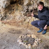 Eine Person in schwarzer Jacke kniet in einer H&ouml;hle vor einem Steinkreis. Sie h&auml;lt einen Stock und l&auml;chelt in die Kamera., &copy; SwabianTravel