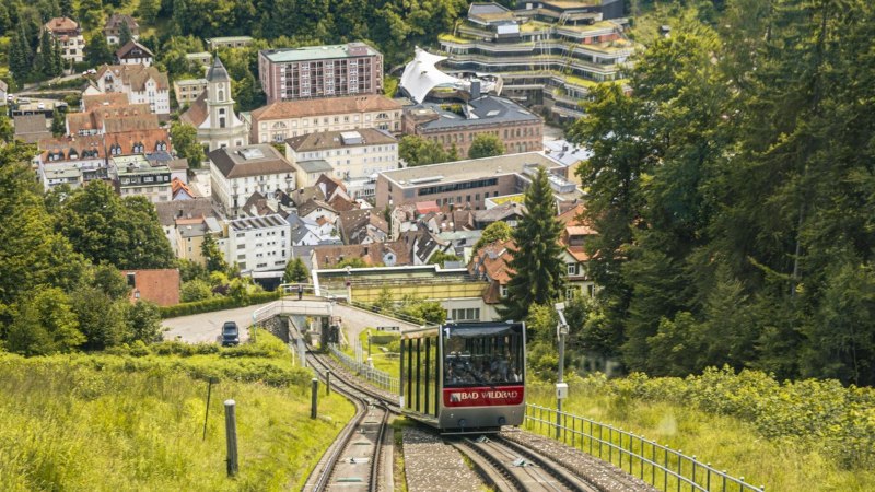 Die Sommerbergbahn fährt durch grüne Landschaft auf Bad Wildbad zu. Im Hintergrund sind Gebäude und Bäume zu sehen., © Stuttgart-Marketing GmbH, Sarah Schmid Die Sommerbergbahn fährt durch grüne Landschaft auf Bad Wildbad zu. Im Hintergrund sind Gebäude und Bäume zu sehen., © Stuttgart-Marketing GmbH, Sarah Schmid