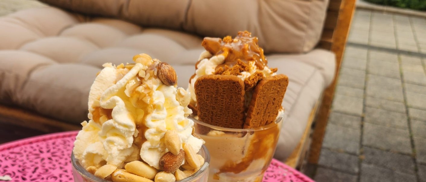 Zwei Eisbecher mit Sahne, Nüssen und Keksen auf einem pinken Tisch im Freien. Im Hintergrund ein beiger Sessel und parkende Autos., © E. Dogan Zwei Eisbecher mit Sahne, Nüssen und Keksen auf einem pinken Tisch im Freien. Im Hintergrund ein beiger Sessel und parkende Autos., © E. Dogan