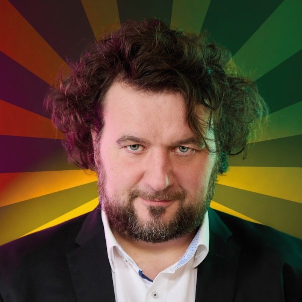 Ein Mann mit lockigem Haar und Bart in einem Anzug vor einem bunten, strahlenförmigen Hintergrund., © Mauerwerk GmbH Ein Mann mit lockigem Haar und Bart in einem Anzug vor einem bunten, strahlenförmigen Hintergrund., © Mauerwerk GmbH