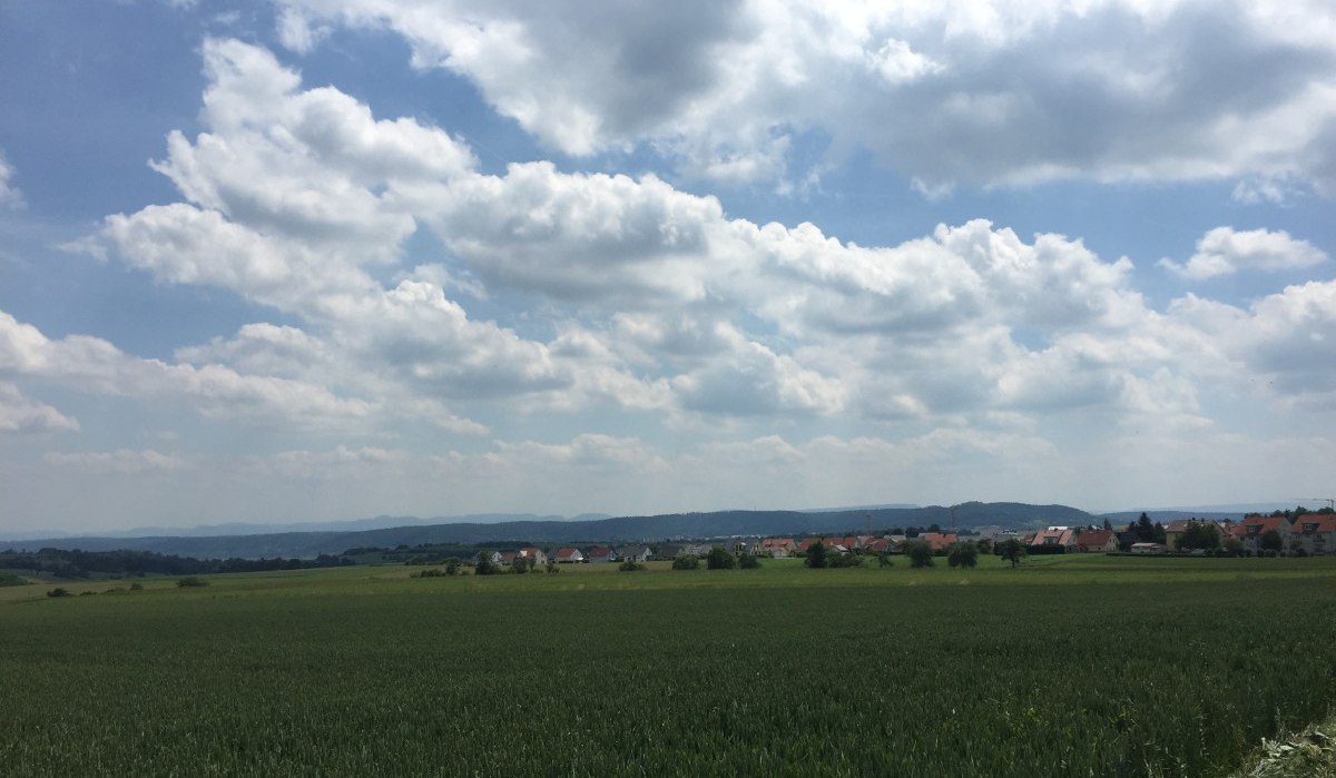 Grüne Felder unter einem bewölkten Himmel, im Hintergrund eine Siedlung vor der Schwäbischen Alb., © www.pro-cycl.de