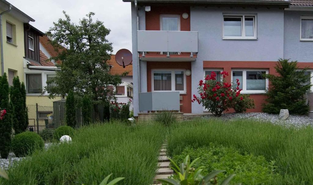 Reihenhaus mit grauem Dach, umgeben von gepflegtem Garten mit grünen Büschen und blühenden Sträuchern. Ein Weg führt durch den Garten., © Nödinger Gastro GmbH