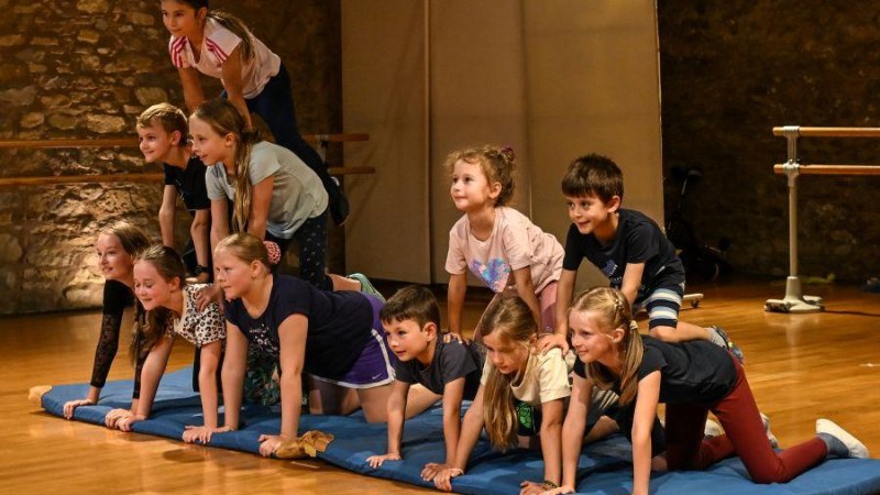 Kinder bauen eine menschliche Pyramide auf einer blauen Matte in einem Raum mit Steinwand und Holzboden., © Kunstschule Labyrinth Kinder bauen eine menschliche Pyramide auf einer blauen Matte in einem Raum mit Steinwand und Holzboden., © Kunstschule Labyrinth