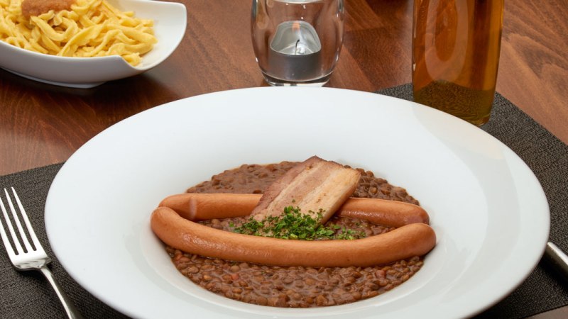 Linseneintopf mit Würstchen und Speck in einem weißen Teller, daneben eine Schüssel Spätzle auf einem Holztisch., © Stuttgart-Marketing GmbH