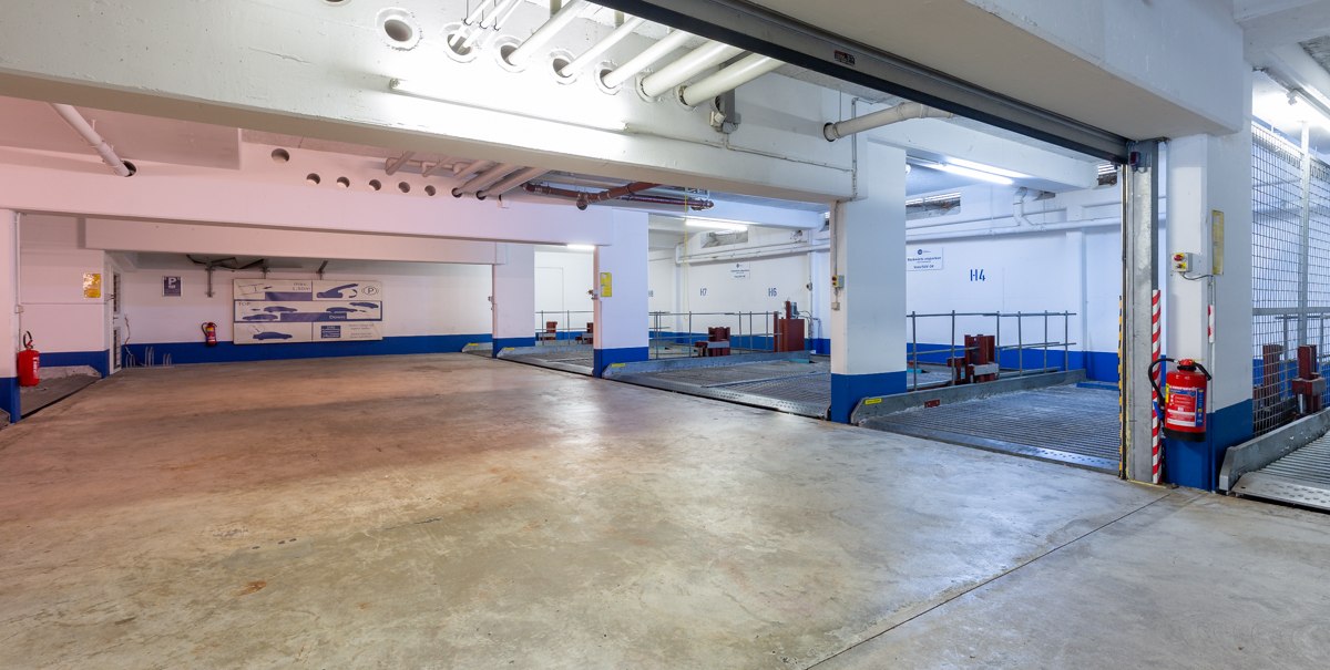Leere Tiefgarage mit markierten Parkplätzen, weißen Wänden und blauen Akzenten. Ein Feuerlöscher ist an der Wand befestigt., © TOMAS Leere Tiefgarage mit markierten Parkplätzen, weißen Wänden und blauen Akzenten. Ein Feuerlöscher ist an der Wand befestigt., © TOMAS