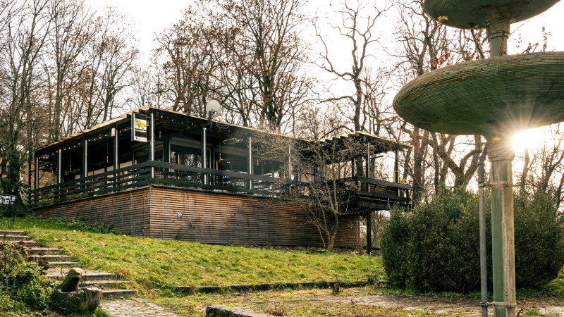 Gebäude mit Holzverkleidung und Terrasse in bewaldeter Umgebung, daneben ein großer, mehrstufiger Brunnen. Sonnenstrahlen scheinen durch die Bäume., © Stuttgart Marketing GmbH, Sarah Schmid Gebäude mit Holzverkleidung und Terrasse in bewaldeter Umgebung, daneben ein großer, mehrstufiger Brunnen. Sonnenstrahlen scheinen durch die Bäume., © Stuttgart Marketing GmbH, Sarah Schmid