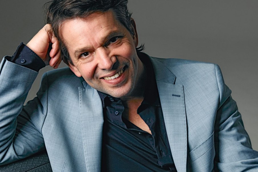 Ein Mann in grauem Anzug lächelt freundlich, den Kopf auf die Hand gestützt. Hintergrund ist neutral grau.