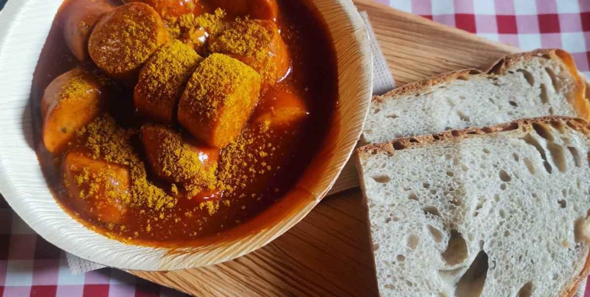 Currywurst in roter Soße mit Curry bestreut, serviert in einer Schale auf einem Holzbrett, daneben zwei Scheiben Brot auf einem rot-weiß karierten Tischtuch., © n/a