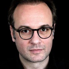 Ein Mann mit Brille blickt direkt in die Kamera. Der Hintergrund ist schwarz, was den Fokus auf das Gesicht lenkt., &copy; links im Bild