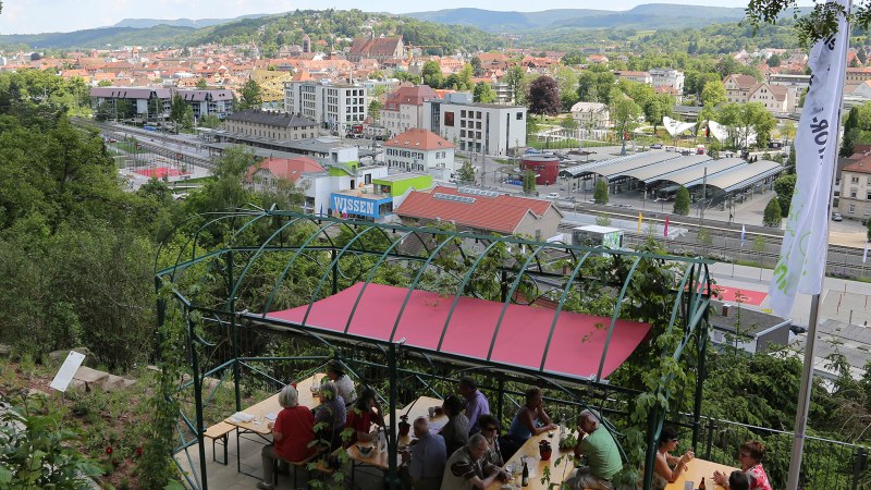 Panoramablick auf eine Stadt mit einem überdachten Sitzbereich im Vordergrund. Menschen sitzen an Tischen, im Hintergrund sind Gebäude und Hügel zu sehen., © Walter Laible