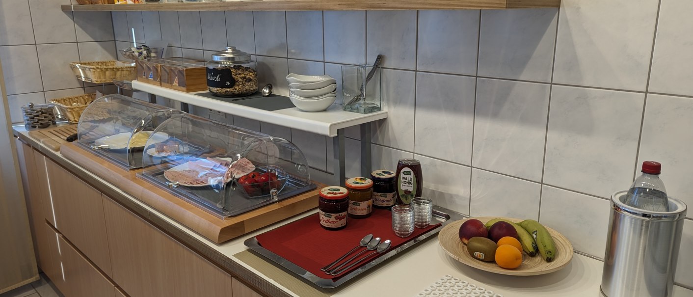 Ein Frühstücksbuffet mit Aufschnitt, Marmeladen, Müsli, Obst und Getränken auf einer Theke in einer Küche., © Gästehaus Ziegler