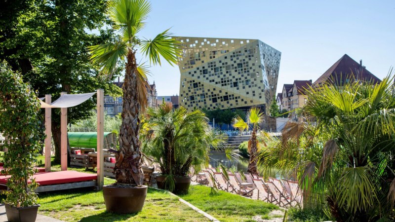 Modernes Geb&auml;ude mit goldener Fassade in Schw&auml;bisch Gm&uuml;nd, umgeben von Palmen und Liegest&uuml;hlen am Wasser., &copy; T&M GmbH Schw&auml;bisch Gm&uuml;nd