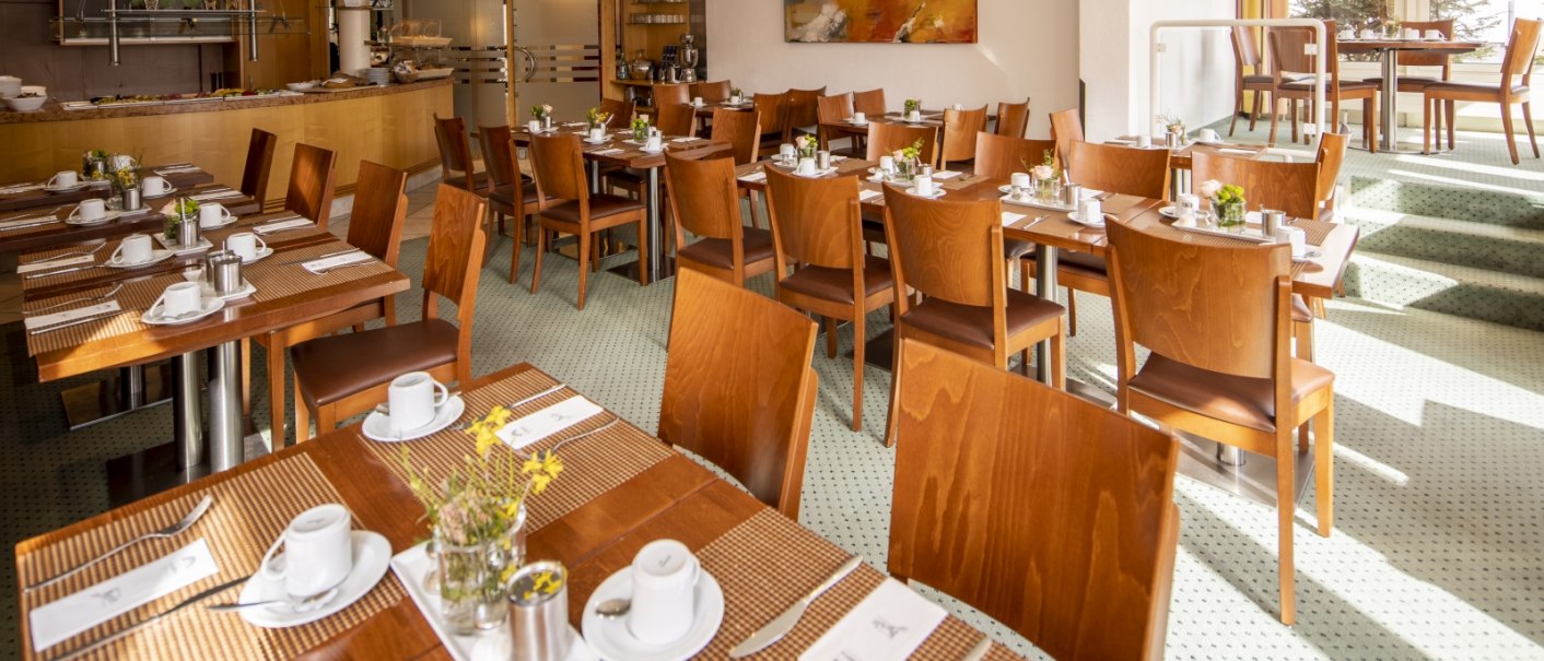 Ein heller Frühstücksraum mit Holztischen und Stühlen, gedeckt mit Geschirr und Blumen. Im Hintergrund ein Buffetbereich., © Hotel Gloria GmbH Restaurant Möhringer Hexle Ein heller Frühstücksraum mit Holztischen und Stühlen, gedeckt mit Geschirr und Blumen. Im Hintergrund ein Buffetbereich., © Hotel Gloria GmbH Restaurant Möhringer Hexle