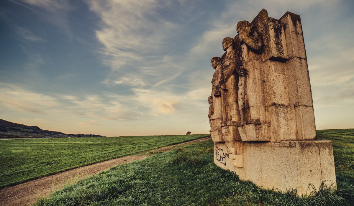 Steinskulptur mit mehreren Figuren auf einem Feldweg. Der Himmel ist leicht bewölkt, die Landschaft weitläufig und grün. Sonnenuntergangsstimmung., © Landratsamt Ostalbkreis