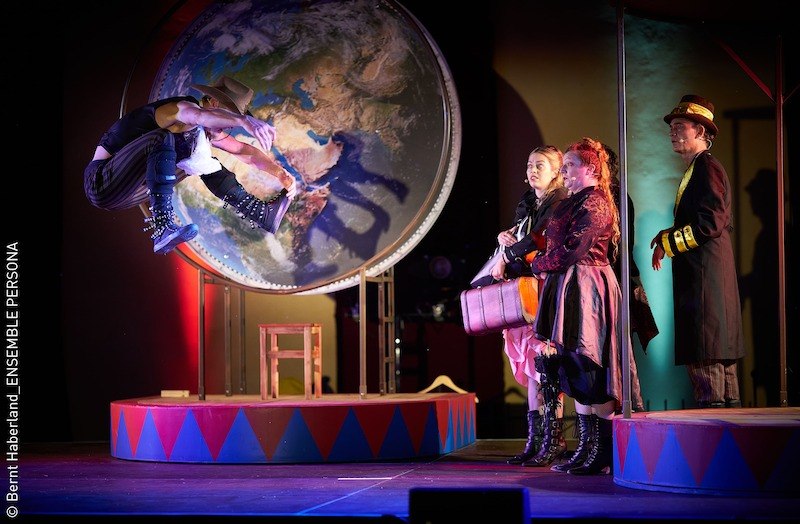 Theaterszene mit einem Akteur, der vor einer großen Weltkarte springt. Drei Personen in Kostümen beobachten ihn. Bühnenbild im Zirkusstil., © Stadt Kornwestheim Kulturmanagement