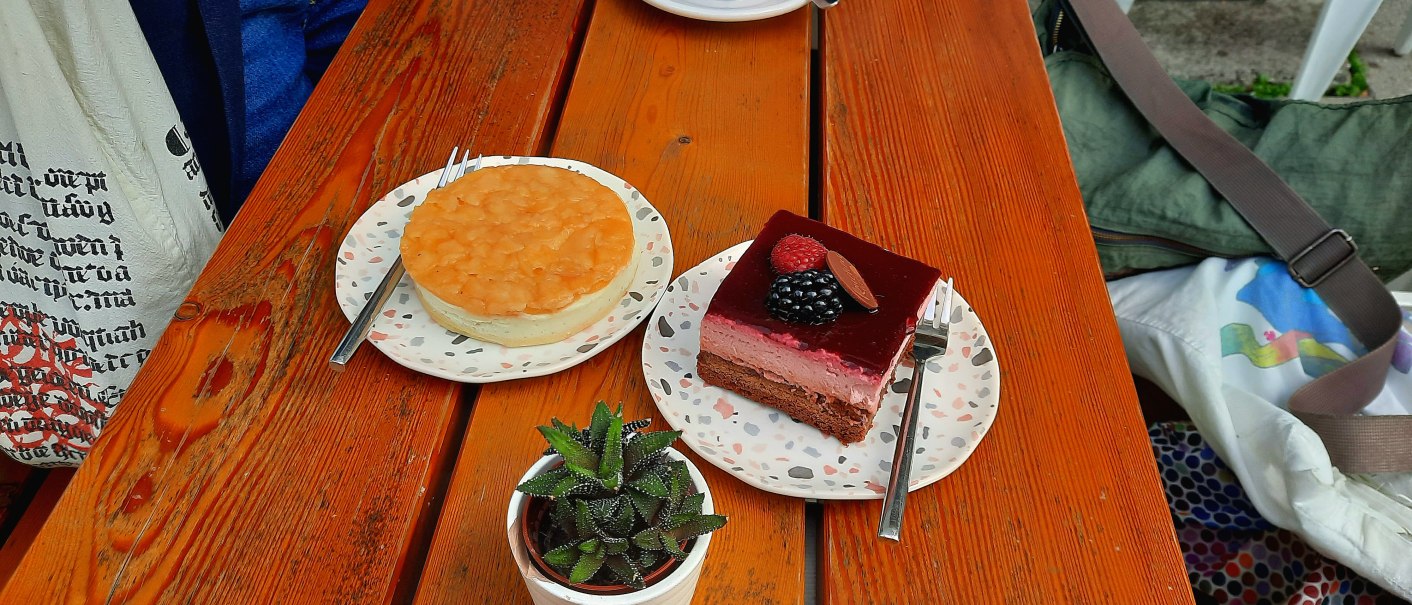 Zwei Kuchenstücke auf Tellern mit Gabeln auf einem Holztisch, daneben eine kleine Pflanze., © SMG