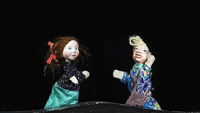 Zwei bunte Handpuppen mit fröhlichen Gesichtern auf einer dunklen Bühne. Eine Puppe hat rote Schleifen im Haar, die andere trägt ein gemustertes Outfit., © Prinzessin Gisela Theater