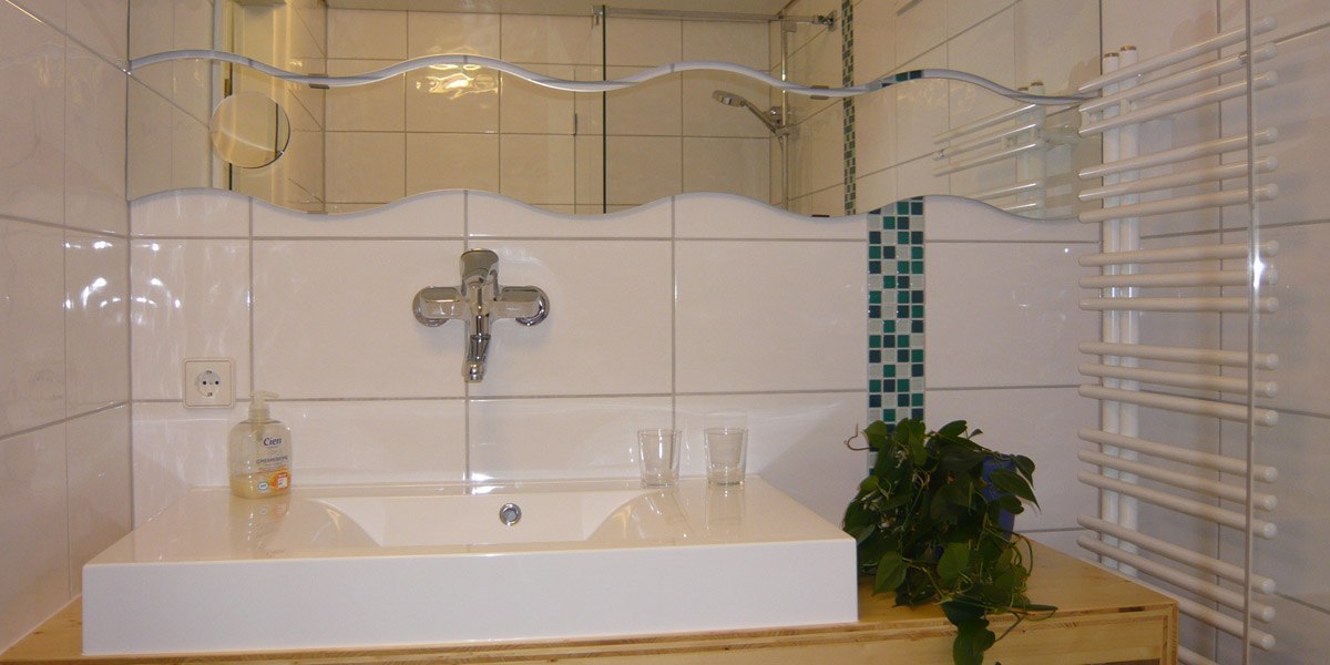 Modernes Badezimmer mit wei&szlig;em Waschbecken, wellenf&ouml;rmigem Spiegel, Handtuchheizk&ouml;rper und gr&uuml;ner Pflanze., &copy; Hotel Hochwald