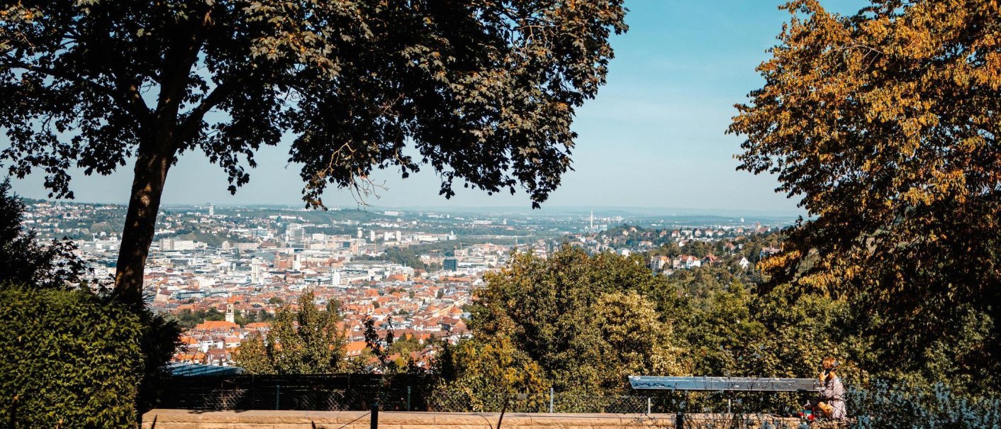Panoramablick auf eine Stadt mit Bäumen im Vordergrund, die den Blick einrahmen. Eine Person sitzt auf einer Bank und genießt die Aussicht., © Stuttgart-Marketing GmbH, Sarah Schmid Panoramablick auf eine Stadt mit Bäumen im Vordergrund, die den Blick einrahmen. Eine Person sitzt auf einer Bank und genießt die Aussicht., © Stuttgart-Marketing GmbH, Sarah Schmid