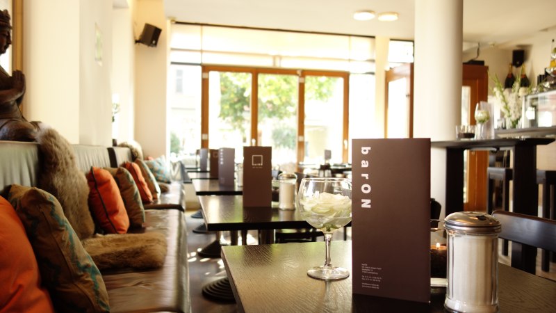 Gemütliches Café mit Kissen auf Bänken, Speisekarten auf Tischen und einem Glas mit Blüten. Große Fenster lassen viel Licht herein., © Andreas Ebert - BARON Gemütliches Café mit Kissen auf Bänken, Speisekarten auf Tischen und einem Glas mit Blüten. Große Fenster lassen viel Licht herein., © Andreas Ebert - BARON