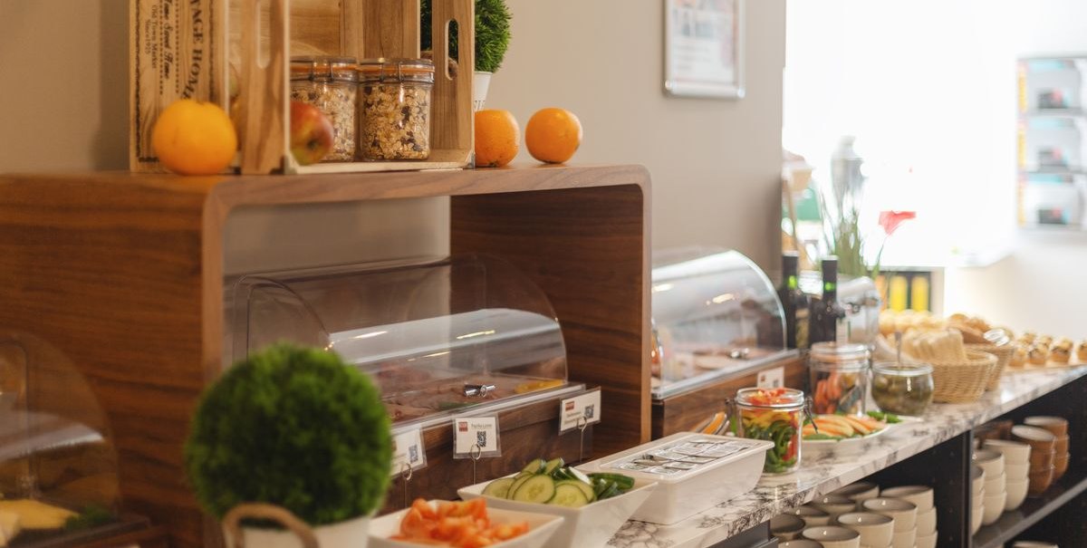 Ein Frühstücksbuffet mit Obst, Gemüse, Müsli und Brotkorb. Holzkisten und Gläser dekorieren den Tisch. Geschirr ist ordentlich gestapelt., © Novum Hospitality
