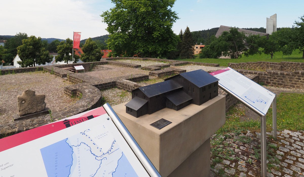 Archäologische Stätte mit Infotafeln und einem Modell eines Gebäudes. Im Hintergrund sind Bäume und moderne Gebäude zu sehen., © T&M GmbH Schwäbisch Gmünd Archäologische Stätte mit Infotafeln und einem Modell eines Gebäudes. Im Hintergrund sind Bäume und moderne Gebäude zu sehen., © T&M GmbH Schwäbisch Gmünd