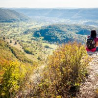 Eine Person sitzt auf einem Hügel und blickt ins Lenninger Tal. Die Landschaft ist von bewaldeten Hügeln und weiten Wiesen geprägt., © hochgehberge