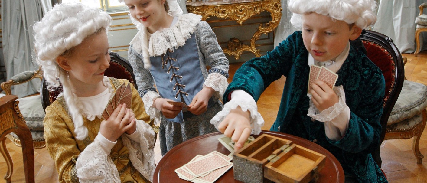 Drei Kinder in historischen Kostümen spielen Karten in einem prunkvollen Raum mit antiken Möbeln und Perücken., © Tourismus und Events Ludwigsburg
