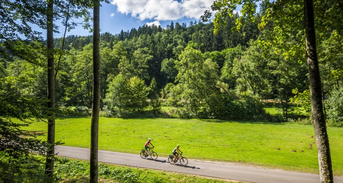 Zwei Radfahrer fahren auf einem asphaltierten Weg durch eine grüne Landschaft mit Wiesen und Bäumen, unter einem blauen Himmel mit weißen Wolken., © Nördlicher Schwarzwald Zwei Radfahrer fahren auf einem asphaltierten Weg durch eine grüne Landschaft mit Wiesen und Bäumen, unter einem blauen Himmel mit weißen Wolken., © Nördlicher Schwarzwald