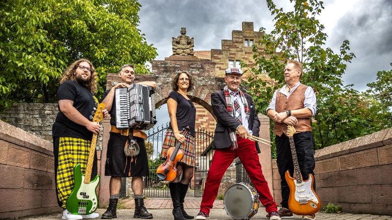 Fünf Musiker posieren mit Instrumenten vor einem alten Torbogen. Sie tragen bunte Kleidung und wirken fröhlich. Der Himmel ist bewölkt., © Backnang