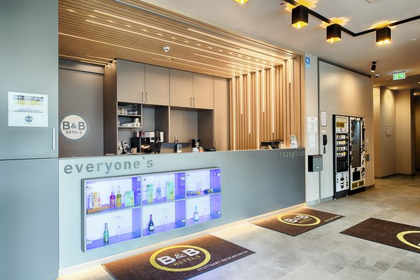 Moderne Hotelrezeption mit beleuchtetem Regal, B&B Hotels Logo und Getr&auml;nkeautomat. Der Empfangsbereich ist hell und einladend gestaltet., &copy; B&B Hotel Stuttgart-Neckarhafen