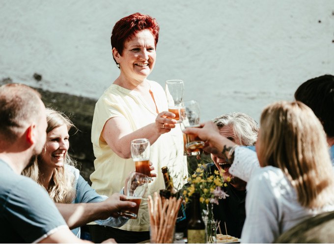 Eine fr&ouml;hliche Gruppe st&ouml;&szlig;t mit Biergl&auml;sern im Freien an. Eine Frau in gelbem Hemd steht und l&auml;chelt. Auf dem Tisch stehen Blumen und Snacks., &copy; Stadt N&uuml;rtingen