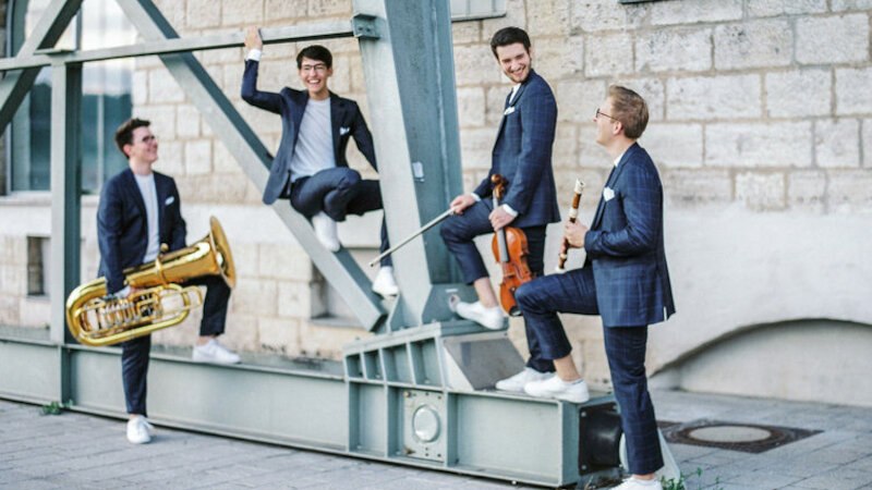 Vier Männer in Anzügen mit Instrumenten posieren lächelnd vor einer Steinmauer. Sie wirken entspannt und fröhlich., © Kammerkonzerte Schwäbisch Hall e.V.