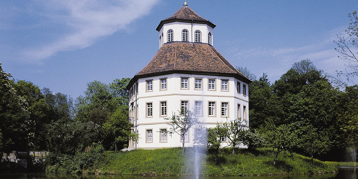 Das Wasserschloss in Oppenweiler mit achteckigem Turm, umgeben von Bäumen. Eine Wasserfontäne sprudelt im Vordergrund., © Stuttgart-Marketing GmbH